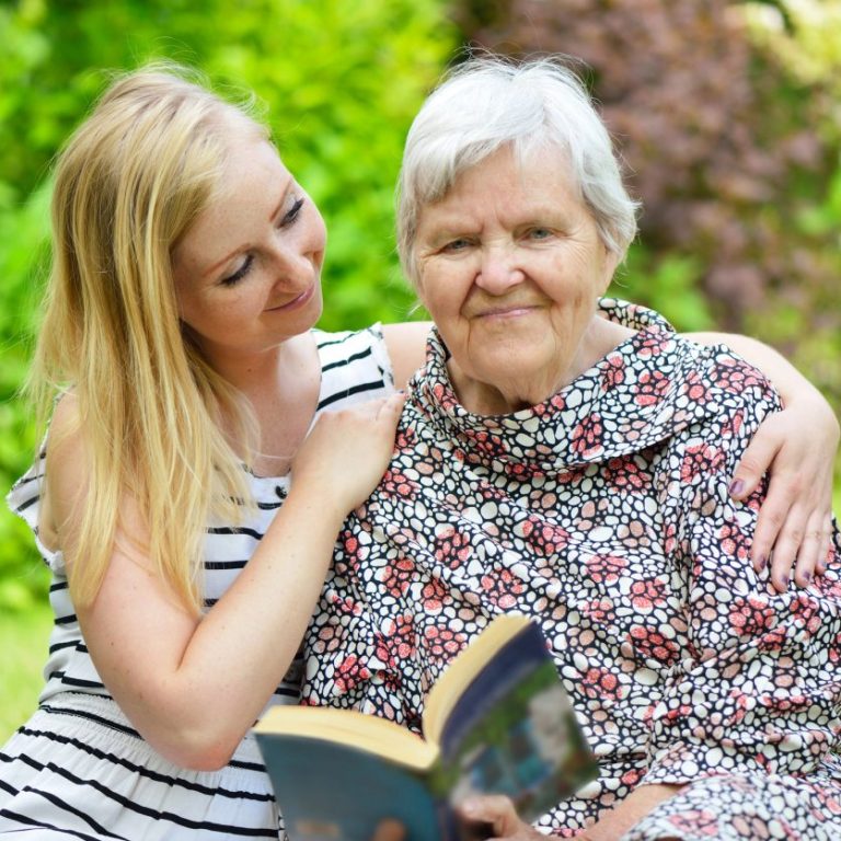 Oma hält Buch in der Hand und schaut lächelnd geradeaus. Tochter umarmt Oma und blickt sie liebevoll an.