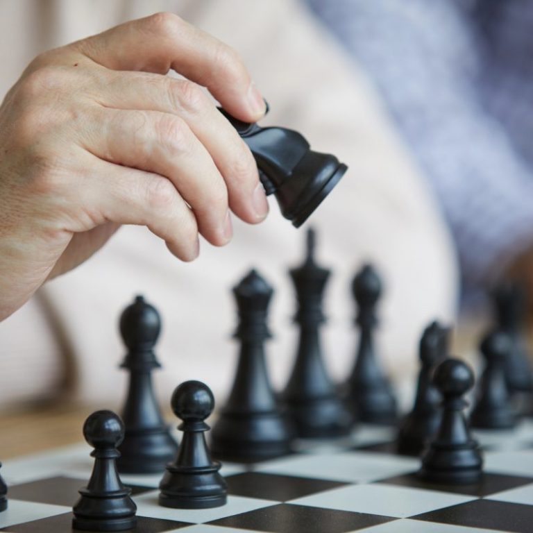 Andreas Bertram Auftragsgedicht Verein Schach Nachaufnahme Schachbrett mit mehreren schwarzen Figuren. Hand hat schwarzen Läufer angehoben um einen Zug auszuführen.